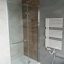 Nowoczesna kabina prysznicowa z przeszklonymi drzwiami, drewnopodobne płytki na ścianie, grzejnik drabinkowy na szarej ścianie, widoczny fragment podłogi z szarych płytek.