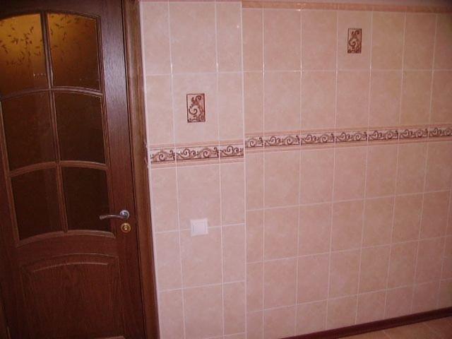 Wykończona ściana w kuchni z beżowych płytek ceramicznych z dekoracyjnym bordiurem i akcentami w postaci kwadratowych płytek z ornamentem, widoczna również drewniana rama drzwi.