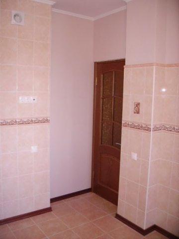 Wykafelkowana kuchnia w trakcie remontu, beżowe płytki na ścianach i podłodze, widoczna instalacja elektryczna i wentylacyjna, drewniane drzwi z szybą