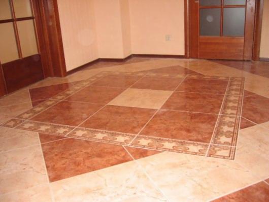 Ułożona podłoga z płytek ceramicznych w dwóch odcieniach brązu, z dekoracyjnym wzorem obramowania w centralnej części, widoczne drzwi z drewnianymi ościeżnicami i fragment ściany w tle.