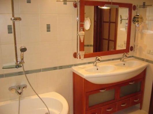 Wykończona łazienka z białą wanną, armaturą chromowaną, umywalką z dwiema bateriami osadzoną na szafce z drewnianymi frontami i dużym lustrem w drewnianej ramie.