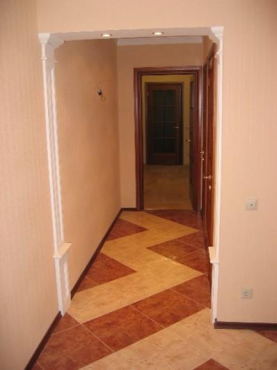 Korytarz z posadzką ułożoną we wzór zygzak z płytek w odcieniach beżu i brązu, widok na otwarte drzwi do dalszej części mieszkania, ściany w kolorze brzoskwiniowym.