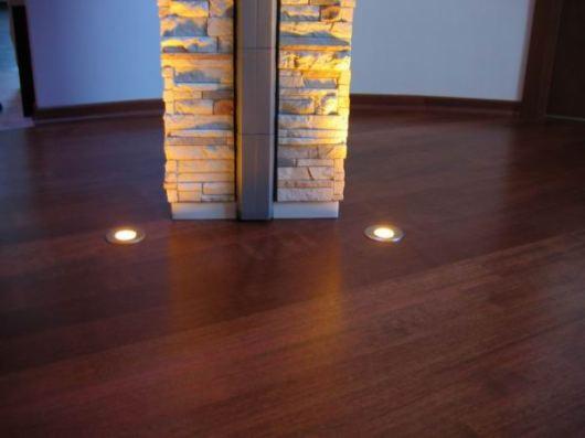 Ciemna, lakierowana drewniana podłoga z wbudowanymi punktowymi lampkami o ciepłej barwie, oświetlającymi kolumnę wykończoną kamieniem dekoracyjnym.