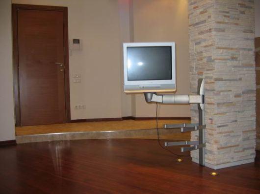 Wykończone wnętrze z ciemną, lakierowaną podłogą, telewizorem na metalowym uchwycie przymocowanym do kamiennej kolumny i drewnianymi drzwiami w tle.