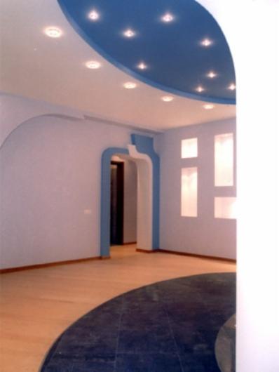 Wykończone wnętrze pokoju z podwieszanym, dwukolorowym sufitem z oświetleniem punktowym, drewnianą podłogą i dekoracyjnymi wnękami w ścianach.