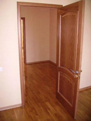 Otwarta, drewniana, dwuskrzydłowa wewnętrzna drzwi z srebrną klamką, widok na parkiet podłogowy i puste pomieszczenie w tle.