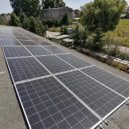 Rząd paneli słonecznych na dachu budynku w Sypniewie, w tle drzewa i zabudowania.