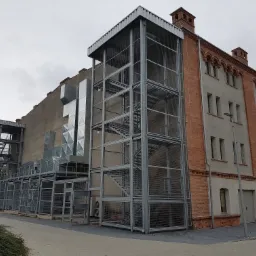 Zewnętrzna, metalowa konstrukcja klatki schodowej z siatką zabezpieczającą, przylegająca do ściany budynku z cegły i szarego tynku, z widocznymi elementami wentylacji na ścianie.