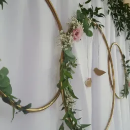 Dekoracja ślubna: złote obręcze z różowymi kwiatami, gipsówką i eukaliptusem na białym tle, z drewnianymi zawieszkami w kształcie ptaków.