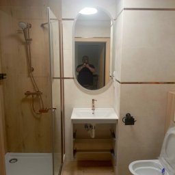 Usługi Remontowo Budowlane - Wykończona łazienka z kabiną prysznicową, umywalką i toaletą w jasnych płytkach. Armatura w kolorze miedzi. W lustrze odbija się osoba robiąca zdjęcie.