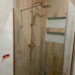 Usługi Remontowo Budowlane - Nowoczesna kabina prysznicowa z deszczownicą w odcieniach brązu i beżu. Widoczne płytki imitujące drewno i wnęka w ścianie. Na ścianie puszka elektryczna.
