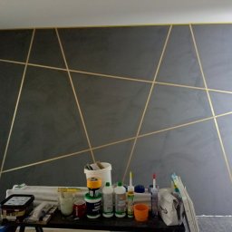 Usługi Remontowo Budowlane - Ściana pomalowana na szaro z geometrycznym wzorem z taśmy malarskiej. Na pierwszym planie narzędzia i materiały malarskie.