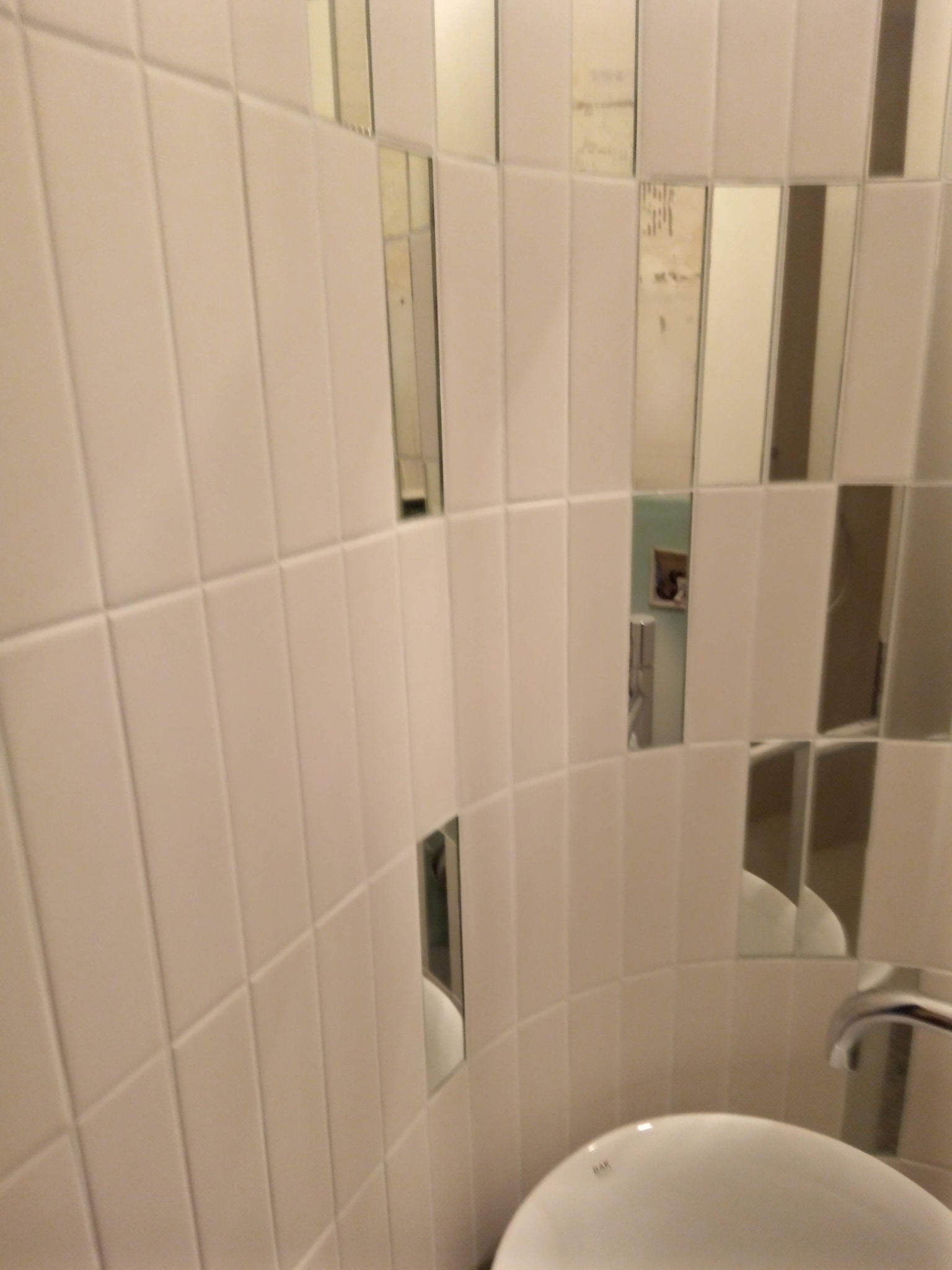 Białe, pionowe płytki z lustrzanymi wstawkami na zaokrąglonej ścianie łazienki, odbijające fragment umywalki i baterii. Nietypowe wykończenie.