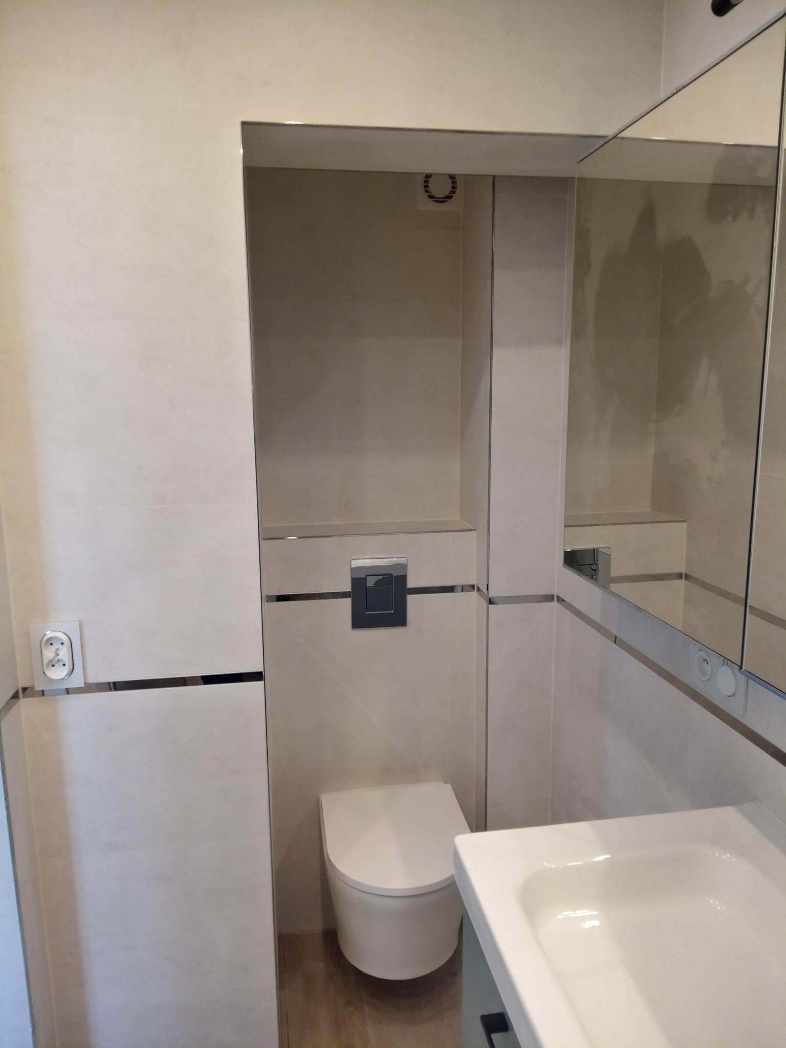 Nowoczesna łazienka w beżowych płytkach z podwieszaną toaletą, umywalką i lustrem. Minimalistyczny design z akcentami chromu. Widoczny fragment wentylacji.