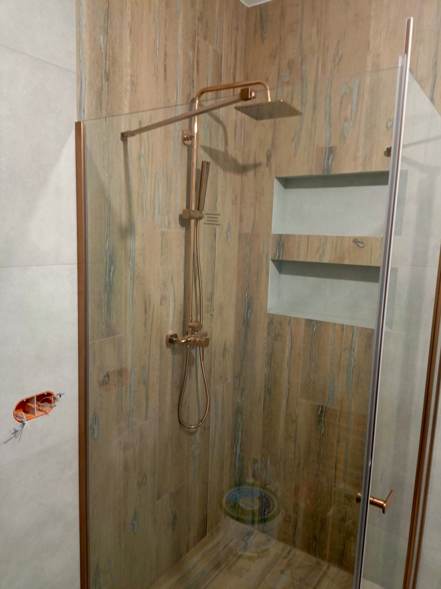 Nowoczesna kabina prysznicowa z deszczownicą w odcieniach brązu i beżu. Widoczne płytki imitujące drewno i wnęka w ścianie. Na ścianie puszka elektryczna.