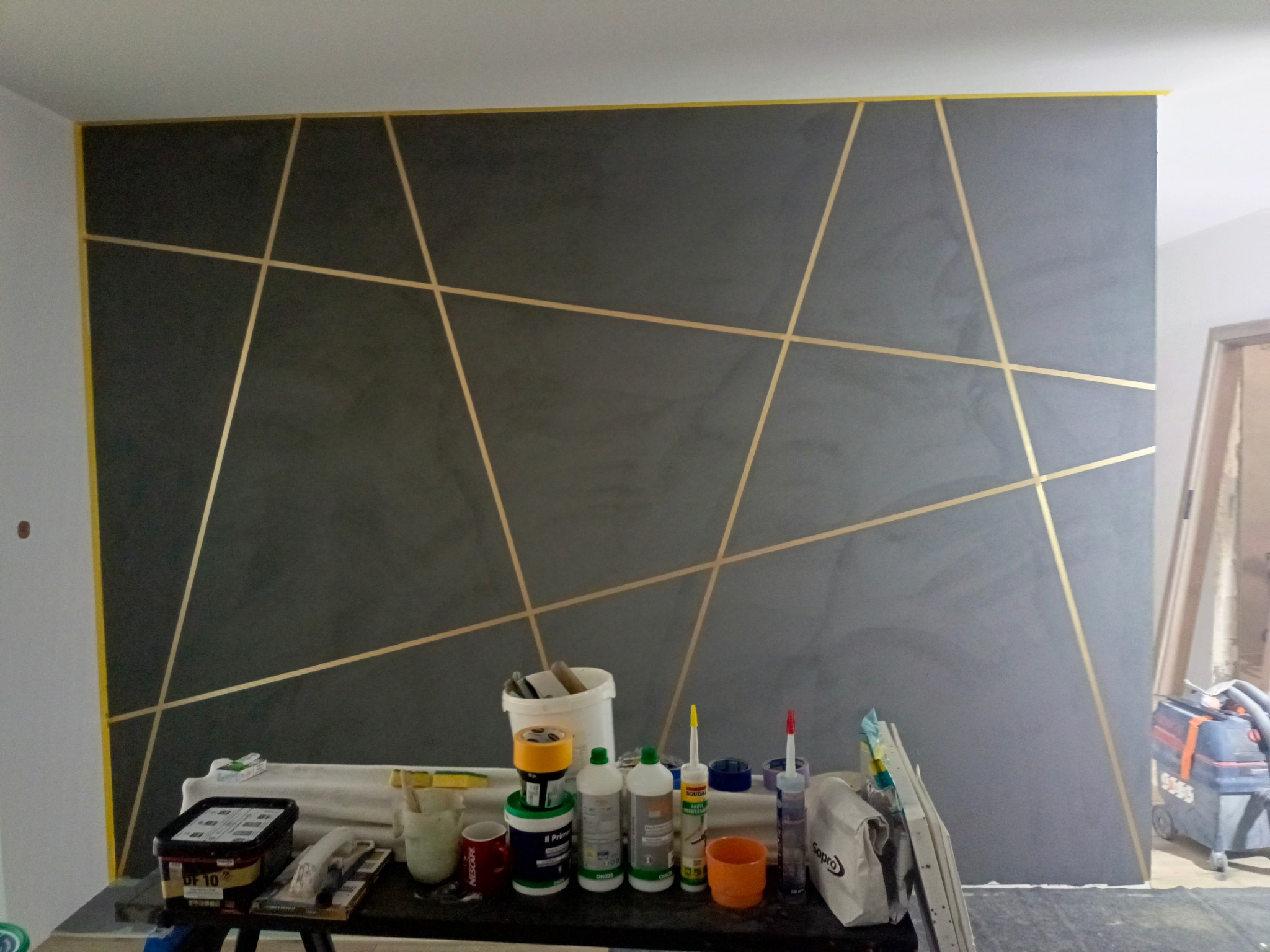Ściana pomalowana na szaro z geometrycznym wzorem z taśmy malarskiej. Na pierwszym planie narzędzia i materiały malarskie.