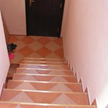 Klatka schodowa z brązowo-beżowymi płytkami w romby, widok z góry na schody prowadzące do ciemnych drzwi. U dołu schodów leżą buty.