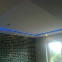 Nowoczesne wnętrze z podwieszanym sufitem, oświetleniem LED, imitacją cegły na ścianie i widokiem na zieleń za oknem. Elementy wykończenia w szarościach i błękicie.