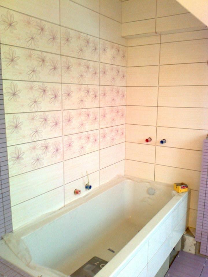 Wanna w rogu łazienki na poddaszu, obłożona jasnymi płytkami z motywem kwiatowym, widoczne wystające rurki i pudełko na brzegu wanny.