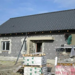 Pracownik na rusztowaniu nakłada warstwę izolacyjną na ścianę zewnętrzną domu jednorodzinnego z szarą dachówką, widoczne okna, drzwi wejściowe i palety z workami zaprawy.