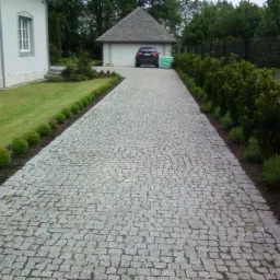 Długa, wybrukowana droga dojazdowa z szarej kostki granitowej, obramowana niskimi, okrągłymi krzewami po lewej i wyższymi, zielonymi krzewami po prawej stronie, prowadząca do garażu z zaparkowanym...