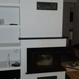 Nowoczesny kominek z czarnymi elementami wykończeniowymi i wnękami na półki po lewej stronie, na podłodze panele.