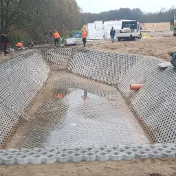 Budowa zbiornika retencyjnego z ażurowych płyt betonowych, widoczni pracownicy w odblaskowych ubraniach i ciężarówka z wywrotką w tle.
