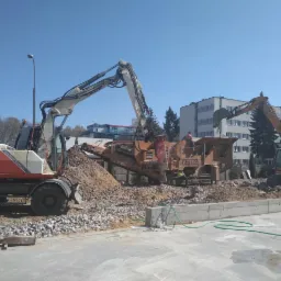 Dwie koparki i kruszarka do gruzu na placu budowy, w tle budynek mieszkalny i błękitne niebo.