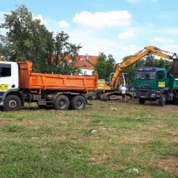 Ż&oacute;łta koparka ładuje ziemię do zielonej ciężar&oacute;wki na trawiastym terenie, w tle biało-pomarańczowa ciężar&oacute;wka i budynki.