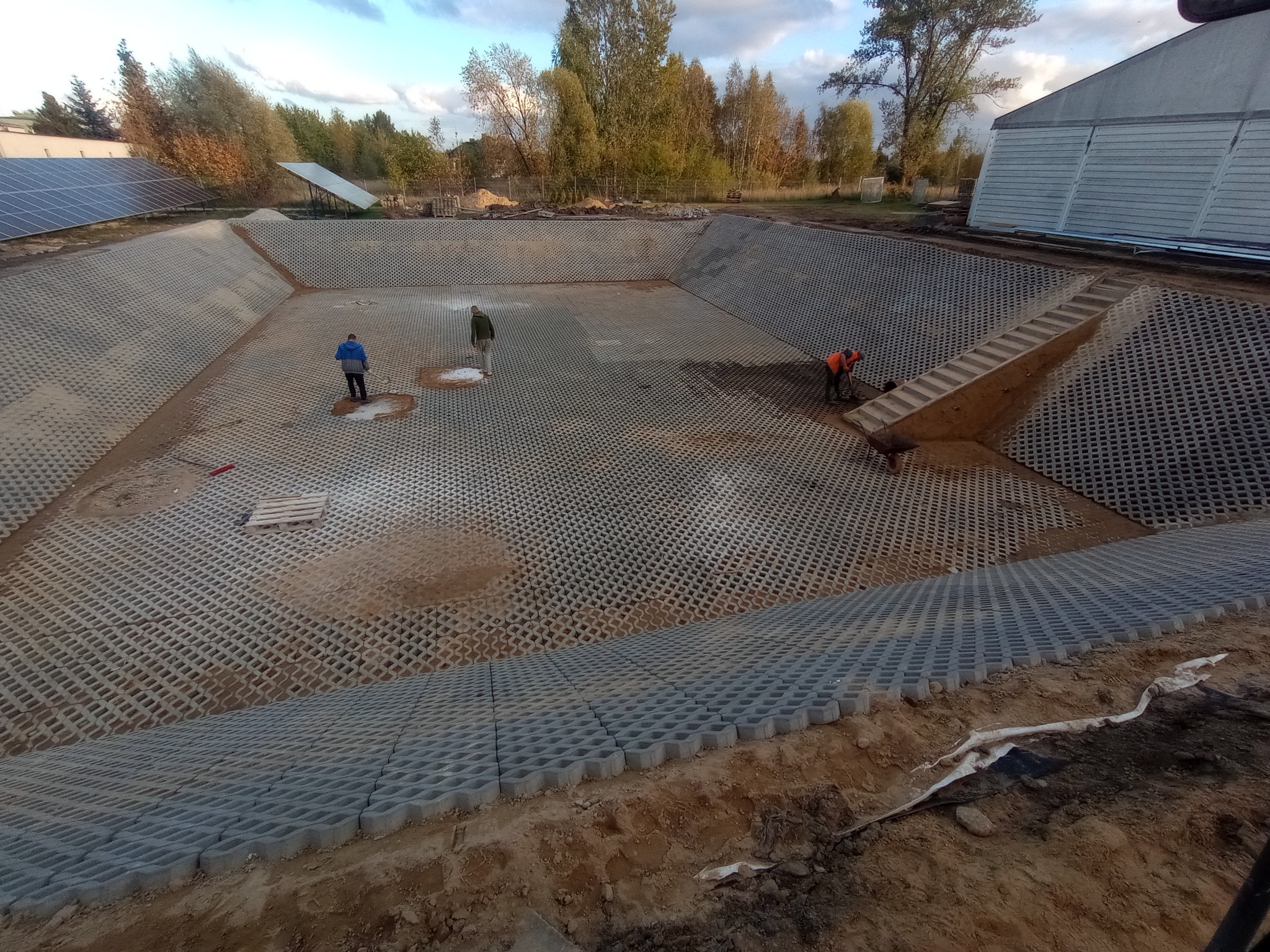 Widok z góry na duży wykop z betonową ażurową kostką na zboczach i dnie, trzech pracowników w oddali, budynek i panele słoneczne w tle.