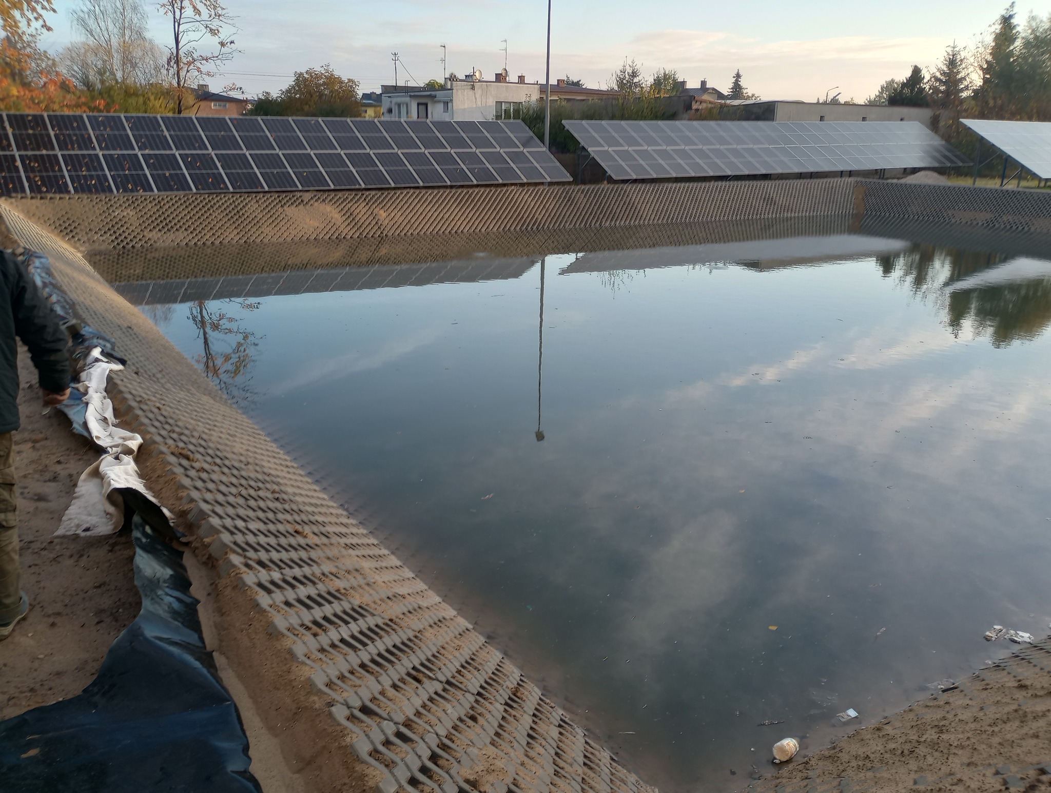 Zbiornik retencyjny z umocnionymi brzegami z ażurowych elementów betonowych, w tle panele słoneczne odbijające się w wodzie, widoczne prace wykończeniowe przy brzegu.