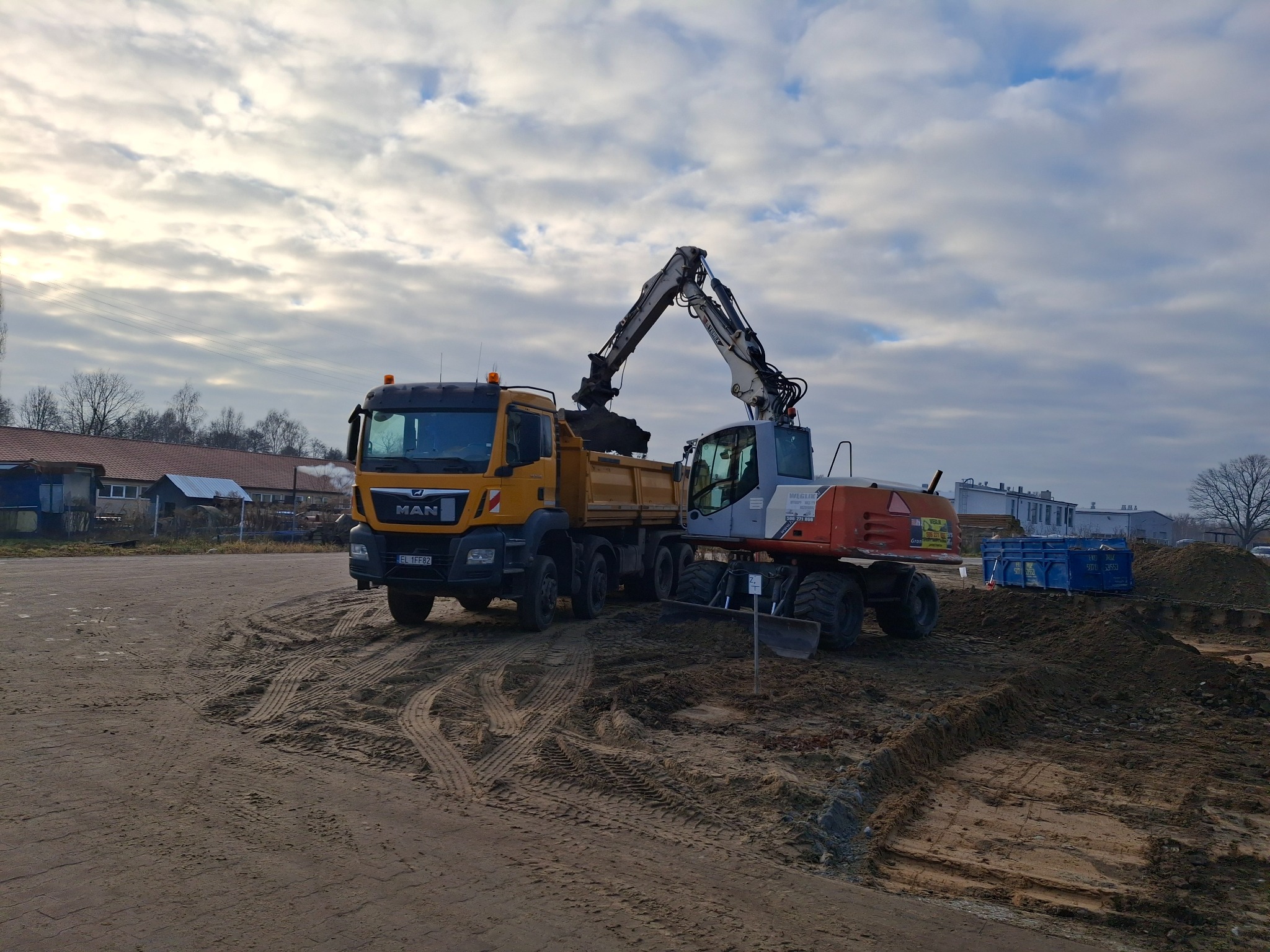 Żółta ciężarówka MAN odbiera ziemię z wykopu za pomocą koparki kołowej na tle pochmurnego nieba.
