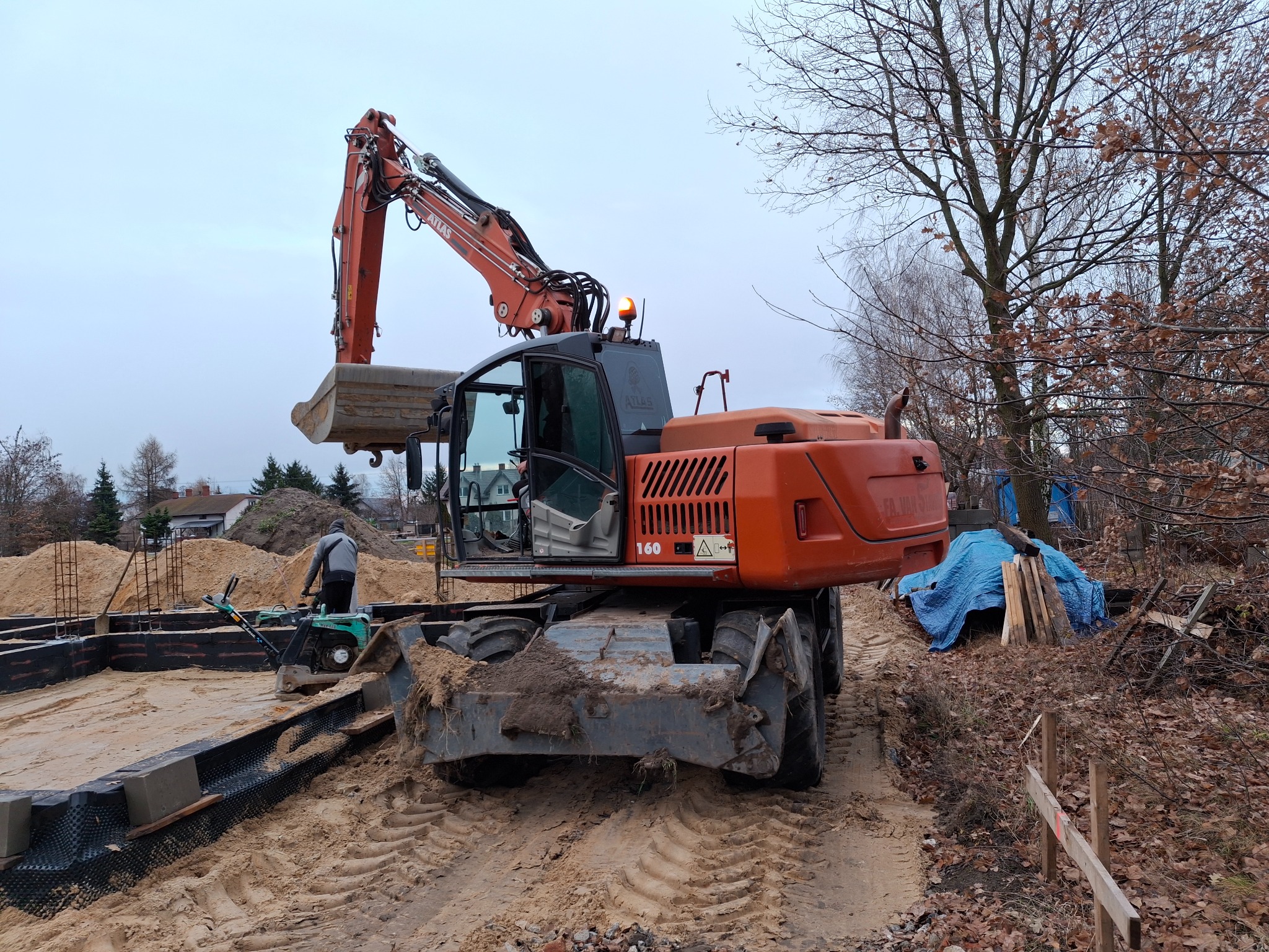Pomarańczowa koparka Atlas 160 z otwartą kabiną na piaszczystym terenie budowy, w tle wykop pod fundamenty, stos piasku i pracownik obsługujący zagęszczarkę.