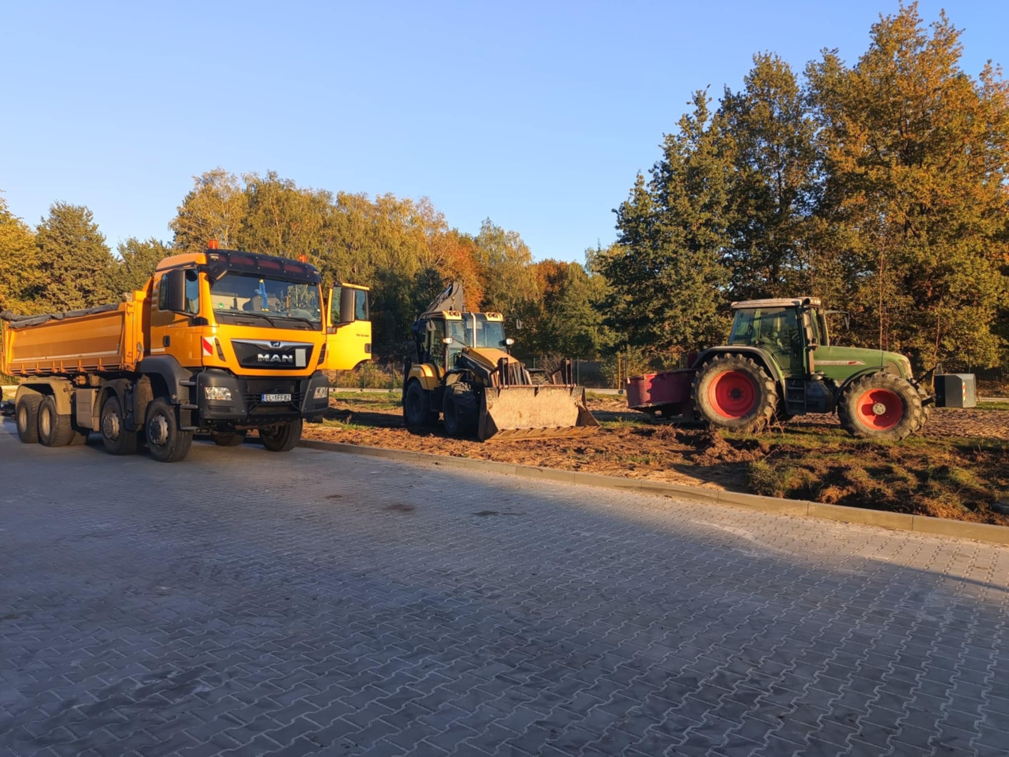 Żółta ciężarówka MAN, żółta koparko-ładowarka i zielony traktor Fendt stoją na nowo ułożonej szarej kostce brukowej, w tle drzewa z jesiennymi liśćmi i błękitne niebo.