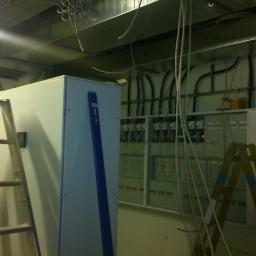 Natel - Rozdzielnica elektryczna w trakcie montażu, widoczne kable, drabina i poziomica, surowy stan wykończenia wnętrza.