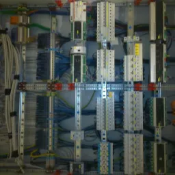 Rozdzielnia elektryczna z licznymi modułami, okablowaniem i szynami montażowymi, widoczne przewody w różnych kolorach, uporządkowane i zamocowane. Elementy zabezpieczone pomarańczowymi klipsami.