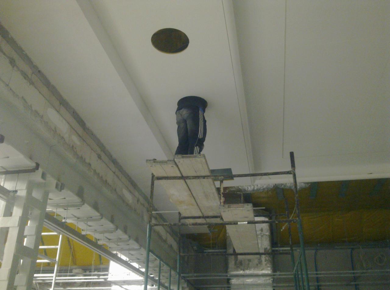 Pracownik na rusztowaniu montuje oświetlenie w suficie podwieszanym, widoczny otwór na lampę.