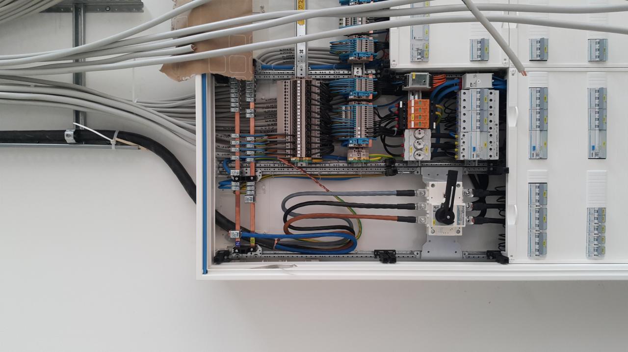 Otwarta skrzynka elektryczna z widocznymi przewodami, bezpiecznikami i innymi elementami instalacji, z przewodami wchodzącymi od góry i boku, w tle widoczne rury instalacyjne.