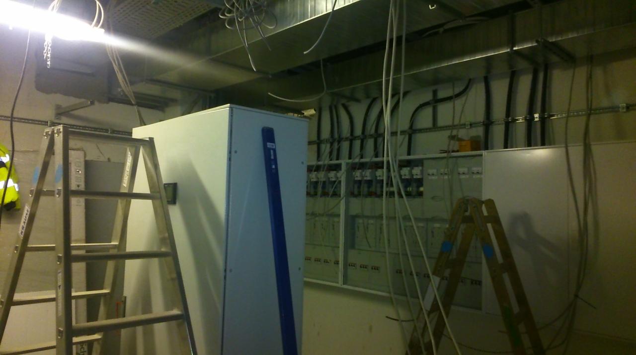 Rozdzielnica elektryczna w trakcie montażu, widoczne kable, drabina i poziomica, surowy stan wykończenia wnętrza.