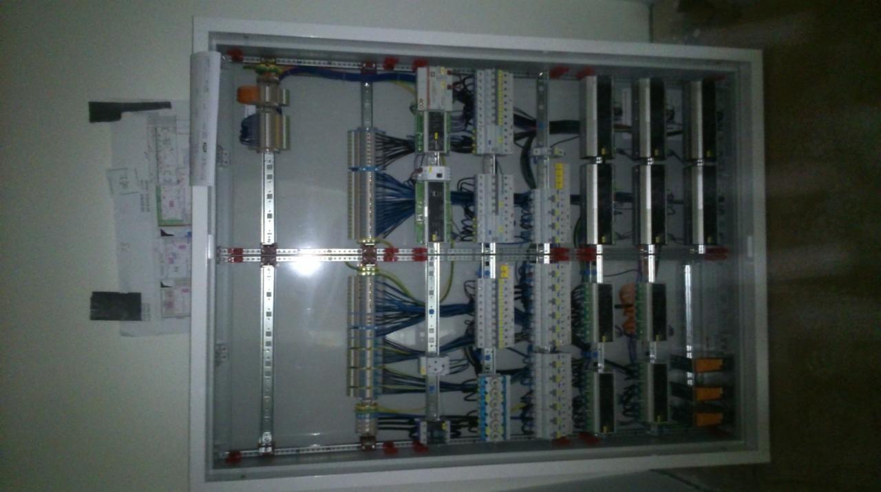 Otwarta skrzynka elektryczna z widocznymi przewodami, bezpiecznikami i elementami montażowymi, schemat instalacji przyklejony obok.