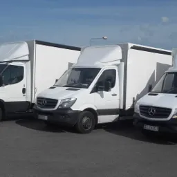 Trzy białe samochody dostawcze zaparkowane obok siebie, pierwszy to Iveco, a dwa kolejne to Mercedesy, z zabudową kontenerową, na parkingu w słoneczny dzień.