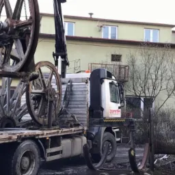 Ciężarówka z dźwigiem transportuje duże, metalowe szpule na kable. Szpule są brudne i wyglądają na używane. Na kabinie ciężarówki widoczne logo 'transtim'. W tle budynek z oknami i drzewa bez liści.
