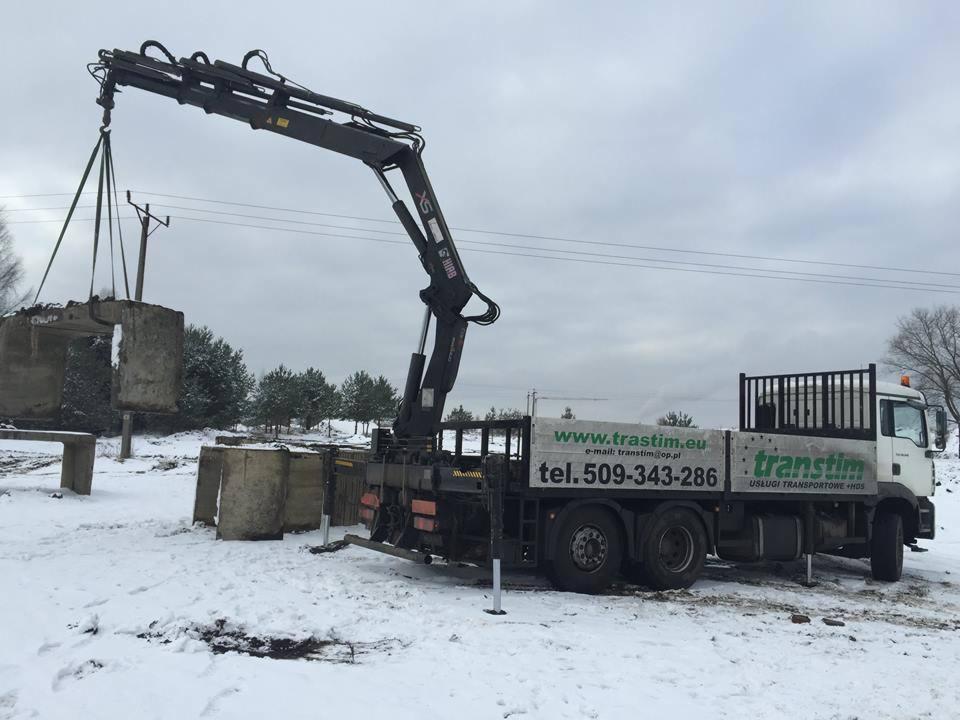 Żuraw samochodowy marki Hiab transportuje betonowy prefabrykat w zimowej scenerii. Widoczny numer telefonu i adres strony internetowej firmy transportowej na burcie ciężarówki.
