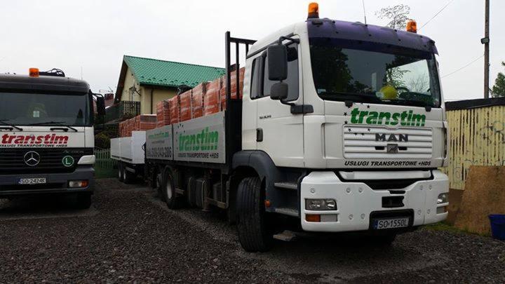 Dwa samochody ciężarowe firmy transportowej z Sosnowca, jeden z przyczepą załadowaną czerwoną cegłą, zaparkowane na żwirowym placu, z widocznym logo firmy na kabinach.