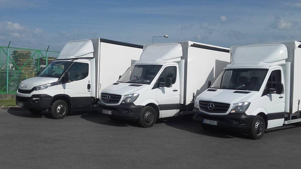 Trzy białe samochody dostawcze zaparkowane obok siebie, pierwszy to Iveco, a dwa kolejne to Mercedesy, z zabudową kontenerową, na parkingu w słoneczny dzień.