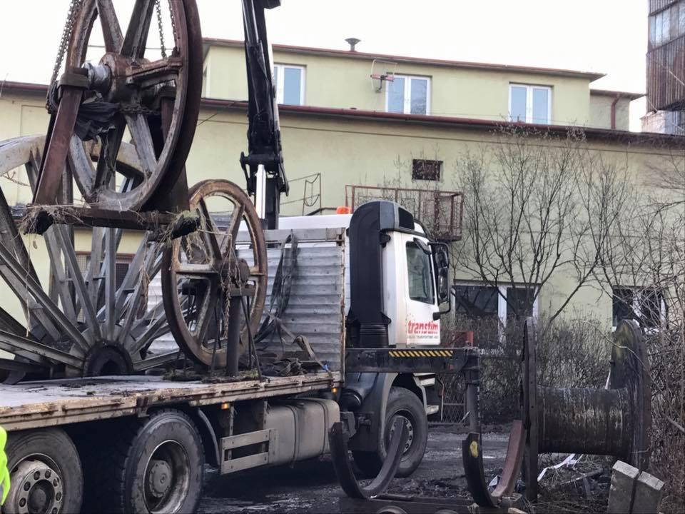 Ciężarówka z dźwigiem transportuje duże, metalowe szpule na kable. Szpule są brudne i wyglądają na używane. Na kabinie ciężarówki widoczne logo 'transtim'. W tle budynek z oknami i drzewa bez liści.