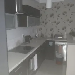 Nowoczesna kuchnia z ciemnymi szafkami, jasnym blatem, płytą indukcyjną i wzorzystą tapetą w tle. Na blacie czajnik elektryczny i akcesoria kuchenne.