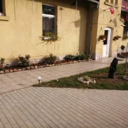 Żółty dom z czerwonym dachem, dekoracjami kwiatowymi w oknach i donicach, oraz ścieżką z kostki brukowej i małym ogródkiem z kamieniami i lampkami.