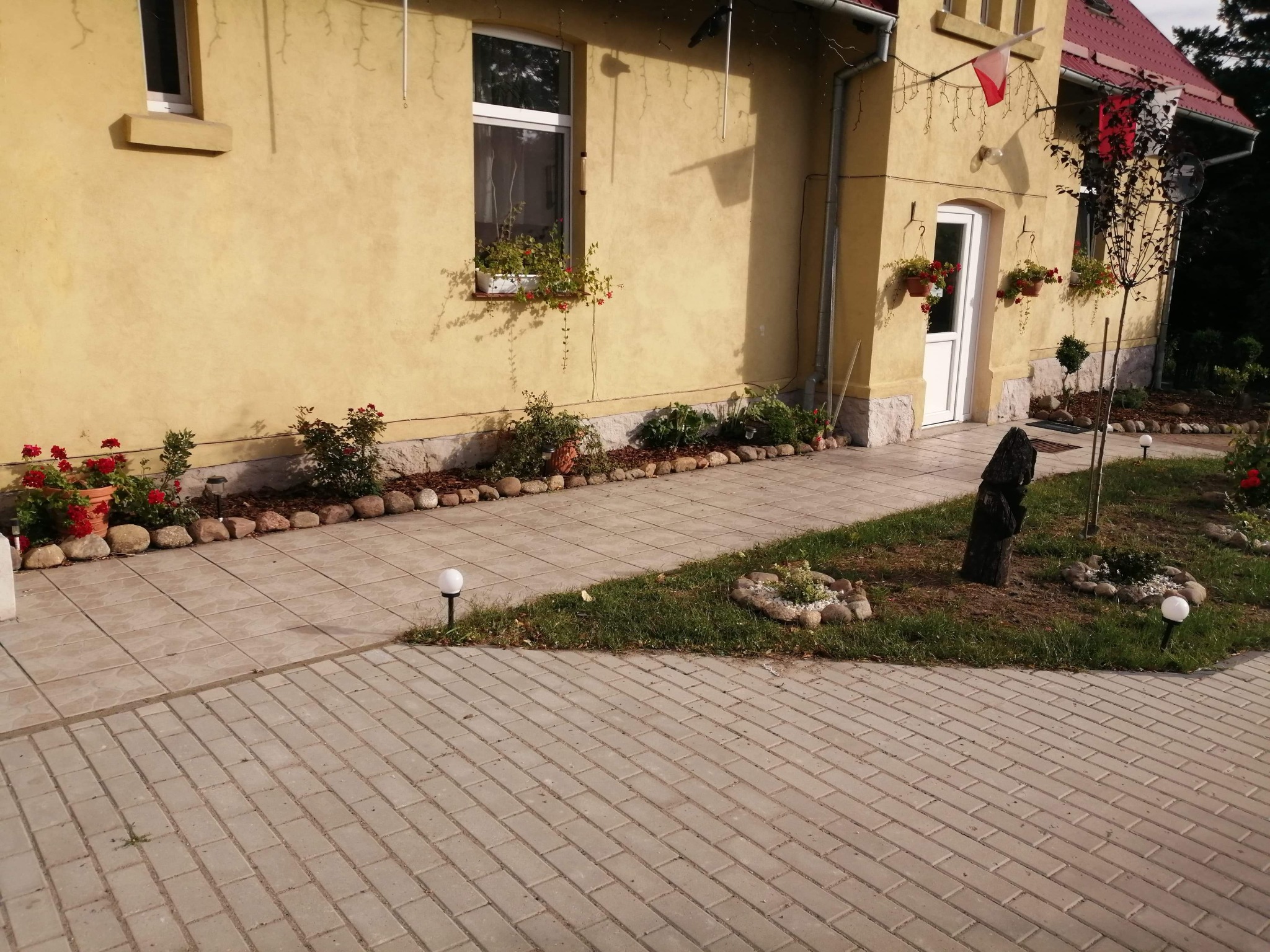 Żółty dom z czerwonym dachem, dekoracjami kwiatowymi w oknach i donicach, oraz ścieżką z kostki brukowej i małym ogródkiem z kamieniami i lampkami.