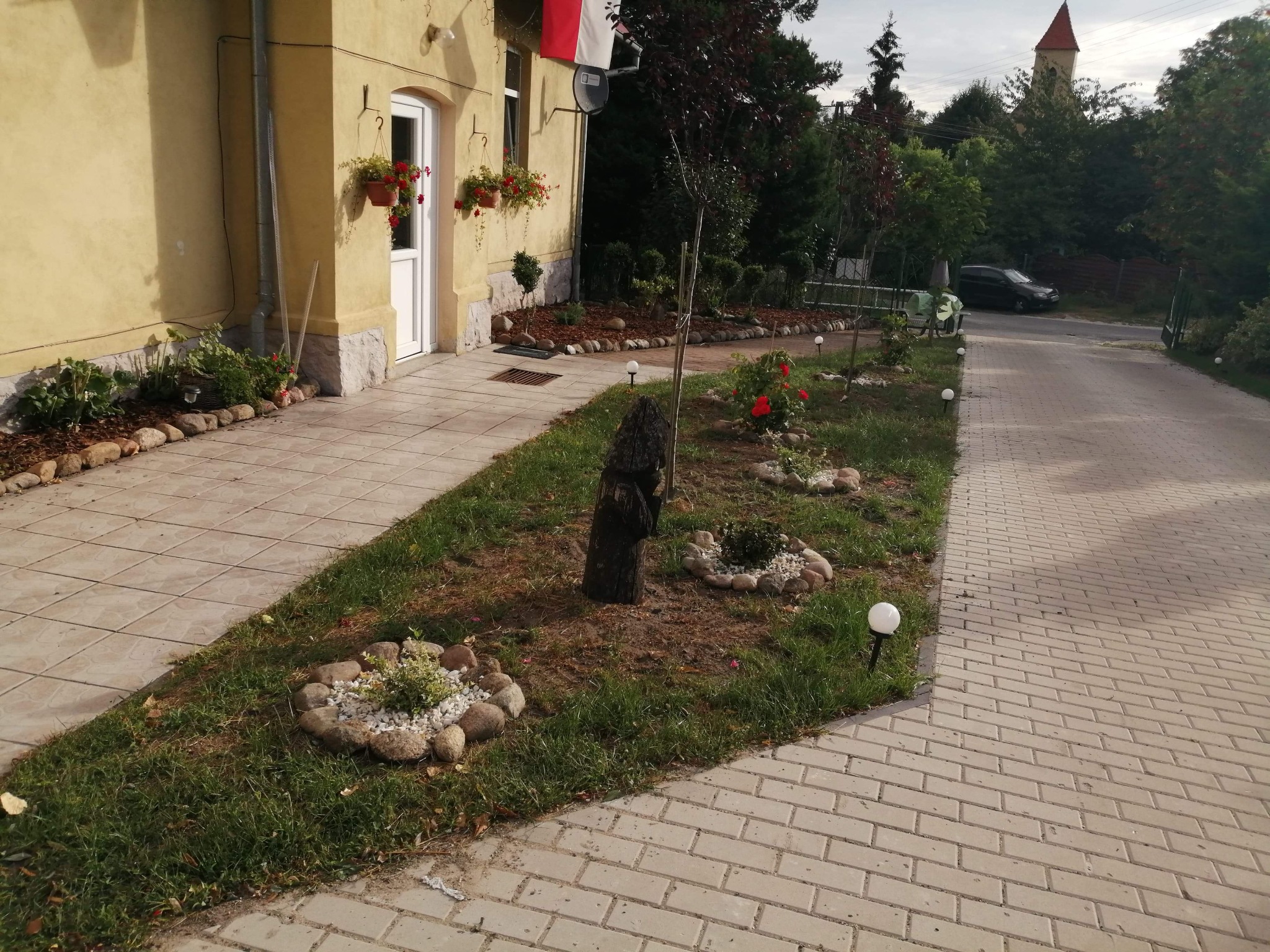Ujęcie z poziomu gruntu na posesję z żółtym budynkiem, kwietnikami pod oknami, zadbanym trawnikiem z rabatami ozdobionymi kamieniami i kulistymi lampami oraz brukowanym podjazdem.
